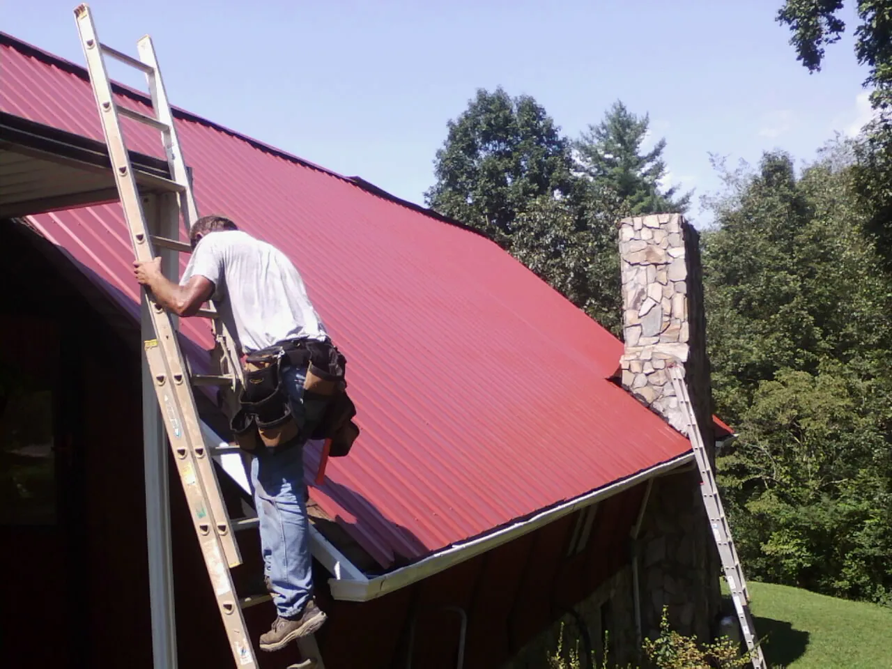 Roofer on a ladder beside a red metal roof and stone chimney — Soffit & Fascia Repair by Roofing Brothers in Whites Creek